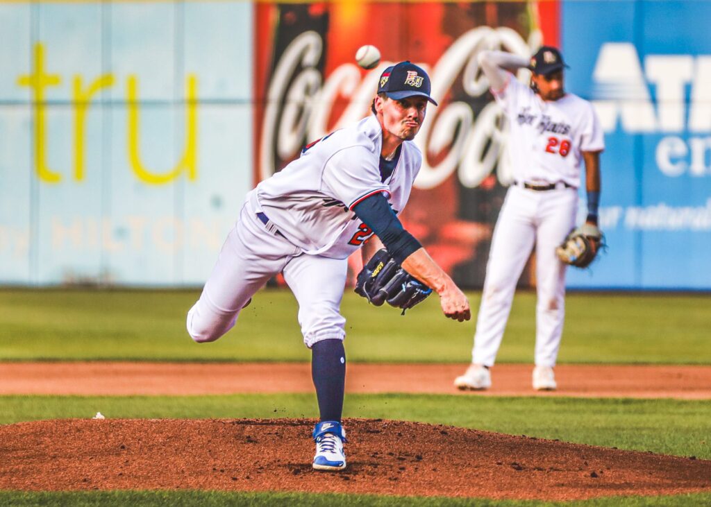 A baseball pitcher in a white uniform, wearing a cap, is in mid-pitch on the mound. Another player stands in the outfield. The background shows advertisements.