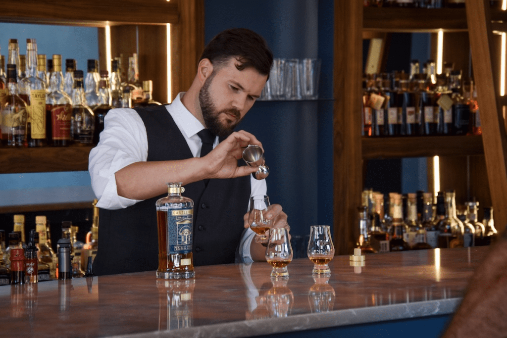 A bartender with a beard and wearing a vest is pouring liquid from a jigger into a glass on a bar counter. Numerous bottles are displayed on shelves in the background.