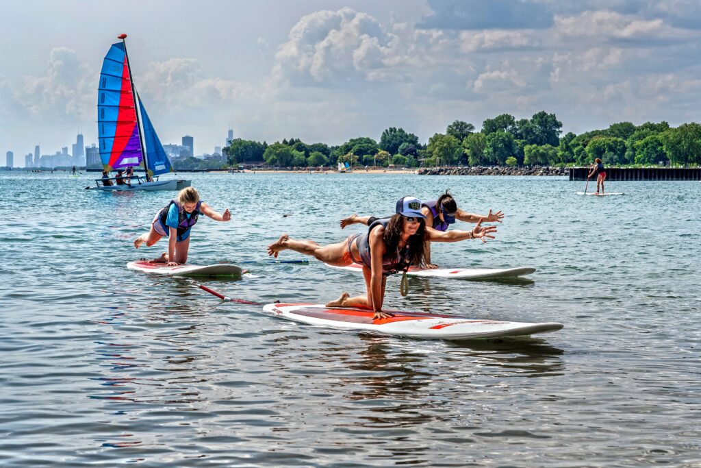 Three people in workout clothes balance on paddleboards in a lake, performing yoga poses. A colorful sailboat and distant city skyline are in the background under a partly cloudy sky.