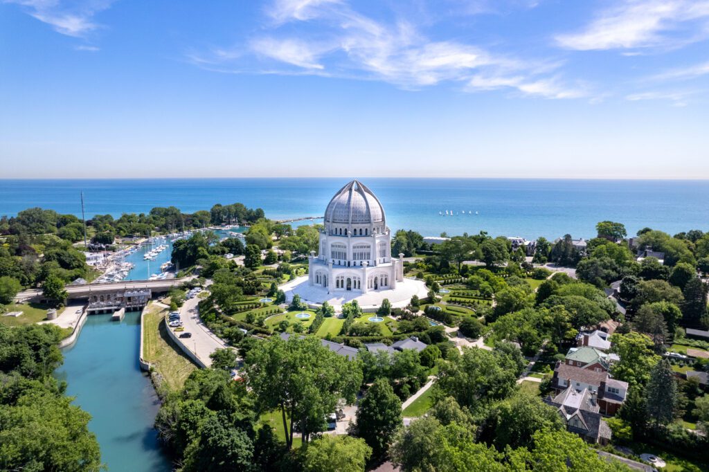 Aerial view of the white Baháí House of Worship with a large dome, surrounded by lush green landscaping. Lake Michigan is visible in the background, along with a small marina and residential buildings. The sky is clear and blue.