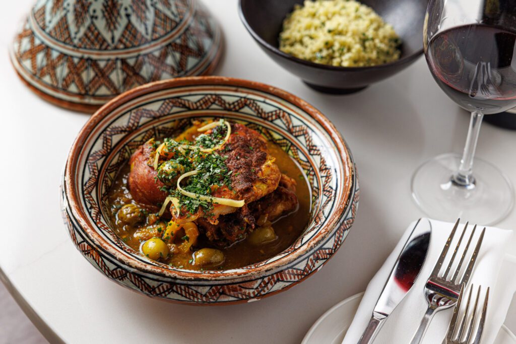 A Moroccan-style dish with spiced meat, olives, and herbs in a decorated bowl. Beside it, theres a covered dish, a bowl of yellow couscous, a glass of red wine, and utensils on a napkin, all set on a white table.