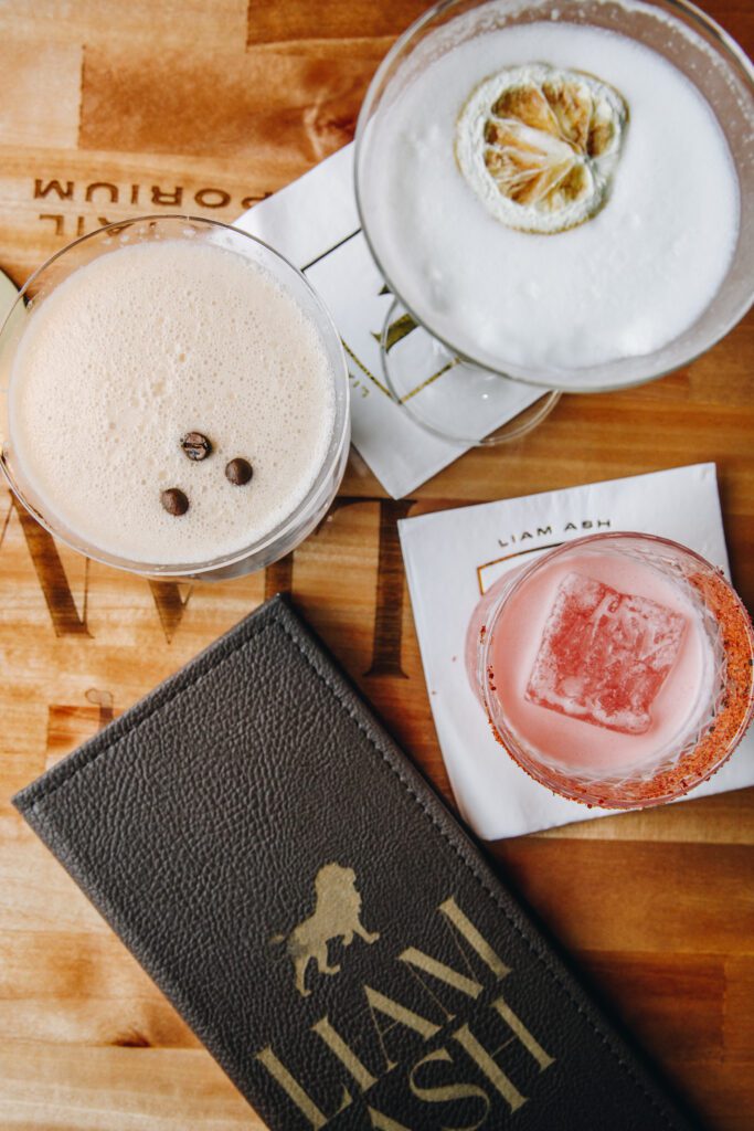 Three cocktails on a wooden table with branded napkins and a menu. One drink is creamy with coffee beans, another has a slice of dried citrus, and the last is pink with a large ice cube. A leather menu with gold text lies nearby.