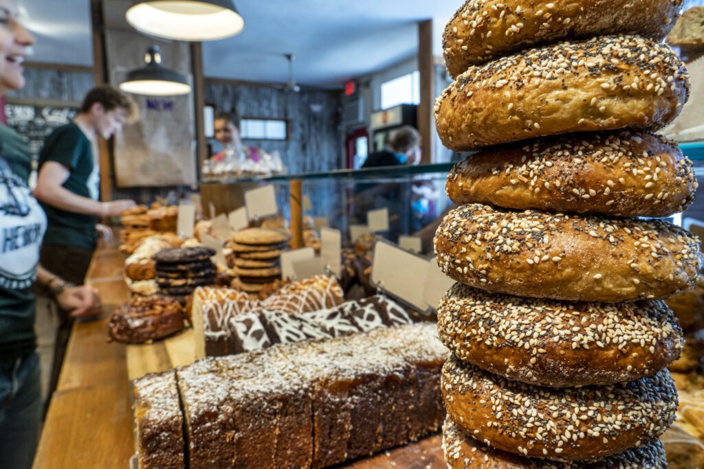 A stack of sesame seed bagels is displayed prominently in a bakery, with various other baked goods like bread and pastries in the background. Customers and staff are visible behind the counter, inside a cozy, rustic shop.