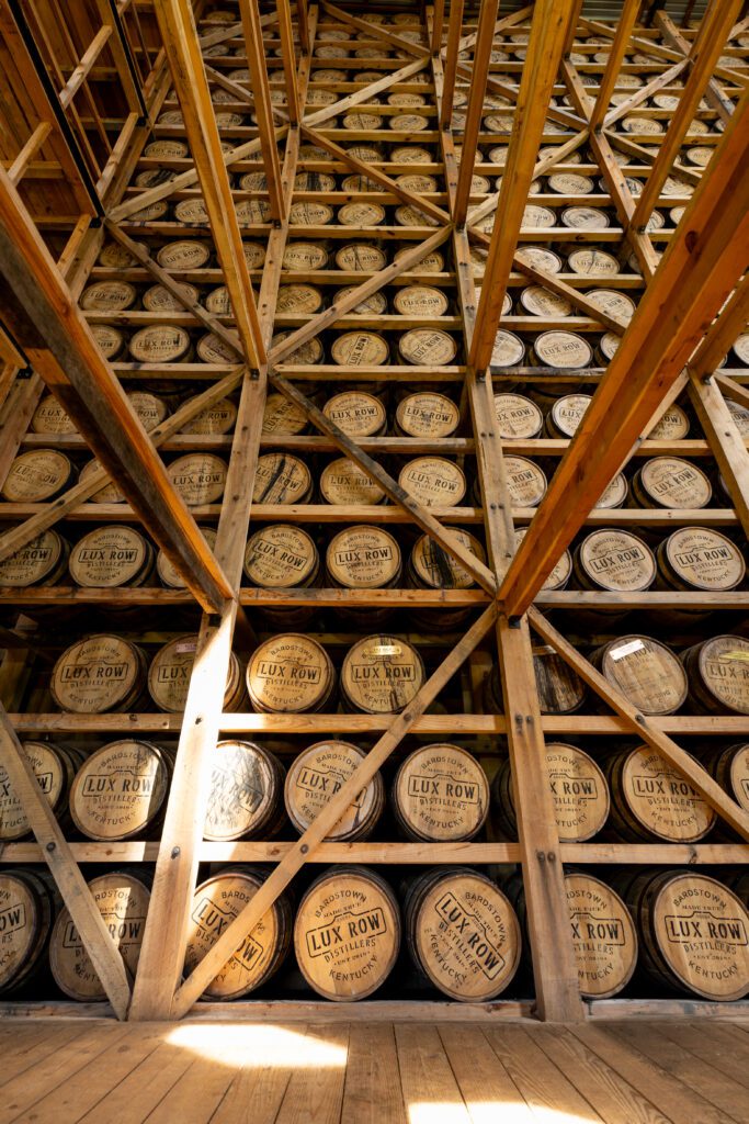 View of stacked wooden barrels in a distillery warehouse. The barrels are arranged on wooden racks, reaching the ceiling, with Lux Row Distillers visible on the barrel tops. Sunlight filters through, creating a warm glow.