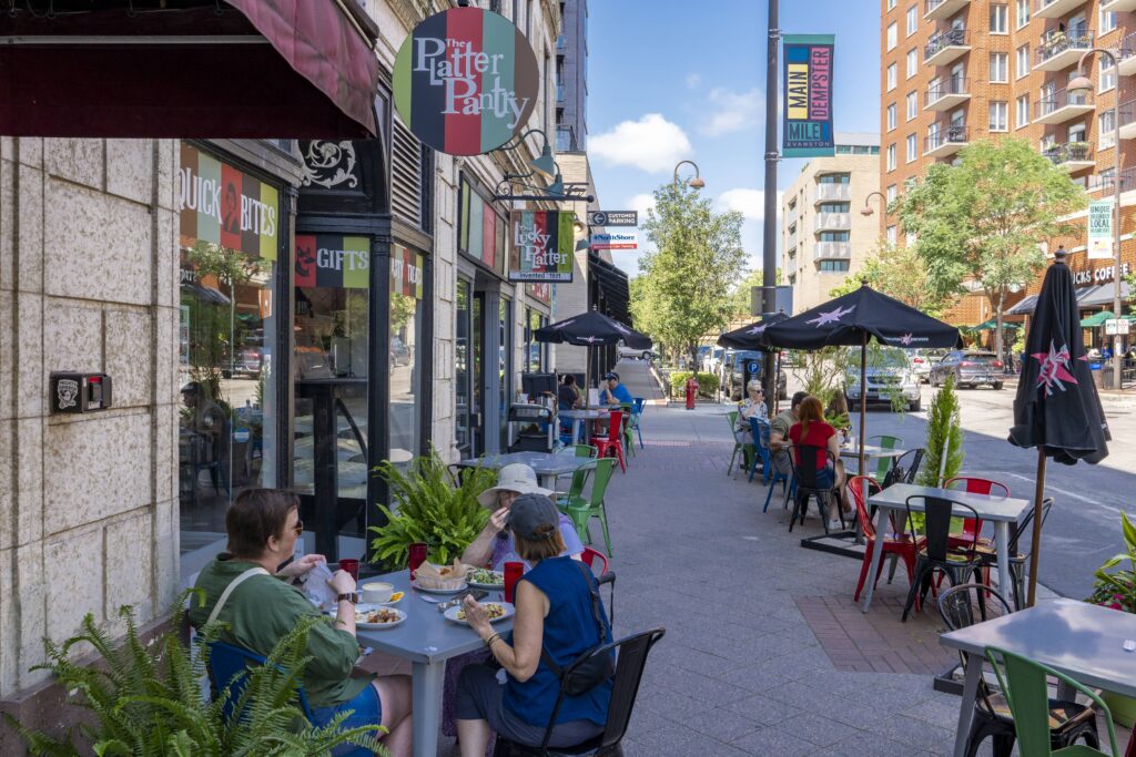 People dine outdoors at tables with blue and red chairs, along a sidewalk cafe on a sunny day. Potted plants and umbrellas provide shade. Nearby, storefronts and a mix of modern and older buildings line the street, with pedestrians in the background.