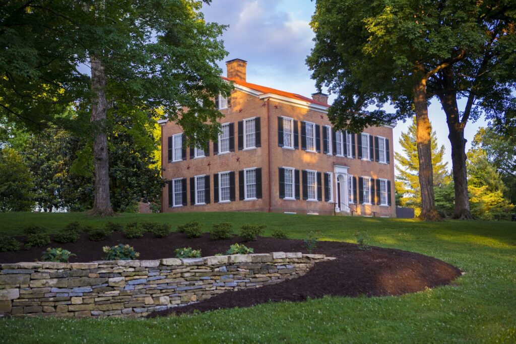 A large, historic brick house with white columns and black shutters sits on a green lawn. A stone retaining wall lines the landscaped garden in the foreground. Tall trees surround the house under a blue sky with clouds.