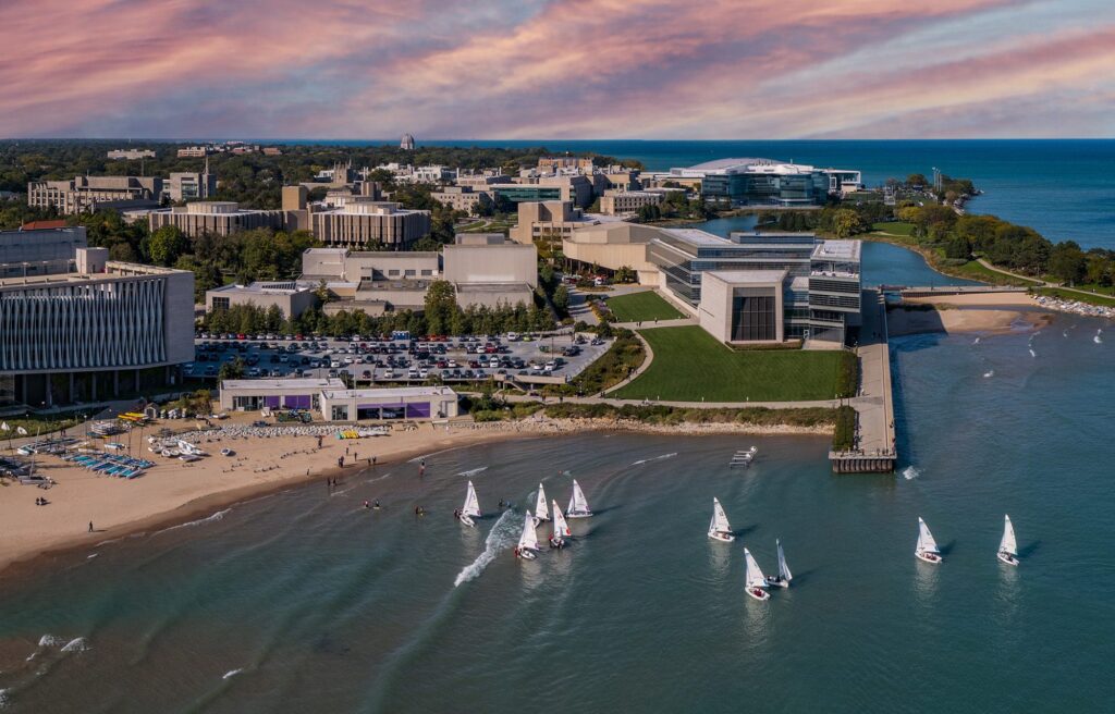 Aerial view of a university campus by a large body of water. White sailboats are clustered near a pier. The campus features modern buildings surrounded by trees, with a sandy beach area visible. The sky is colorful with hues of pink and orange.