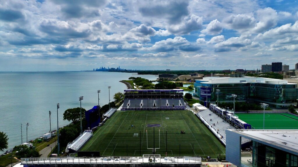 Aerial view of a large sports field near a lake, with bleachers on both sides. In the background, a city skyline is visible under a partly cloudy sky. The area is surrounded by modern buildings and greenery.