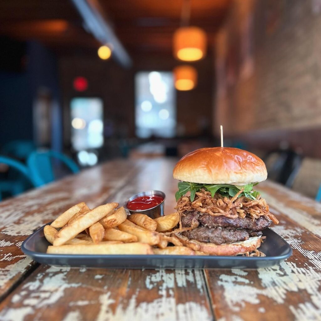 A gourmet burger topped with crispy onions and greens sits on a plate next to a side of thick-cut fries and a small dish of ketchup. The setting is a rustic restaurant with wood tables and low lighting.