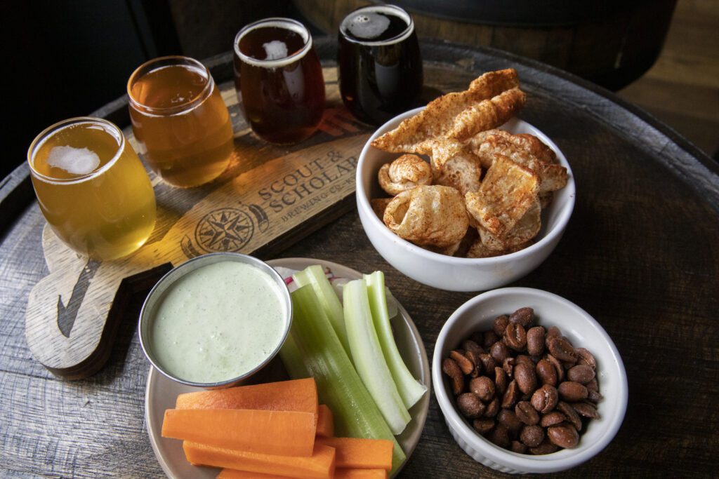 A wooden table displays a beer flight with four glasses of different beers. Nearby, there are bowls with pork rinds and roasted nuts, plus a plate of carrot and celery sticks with a creamy dip.