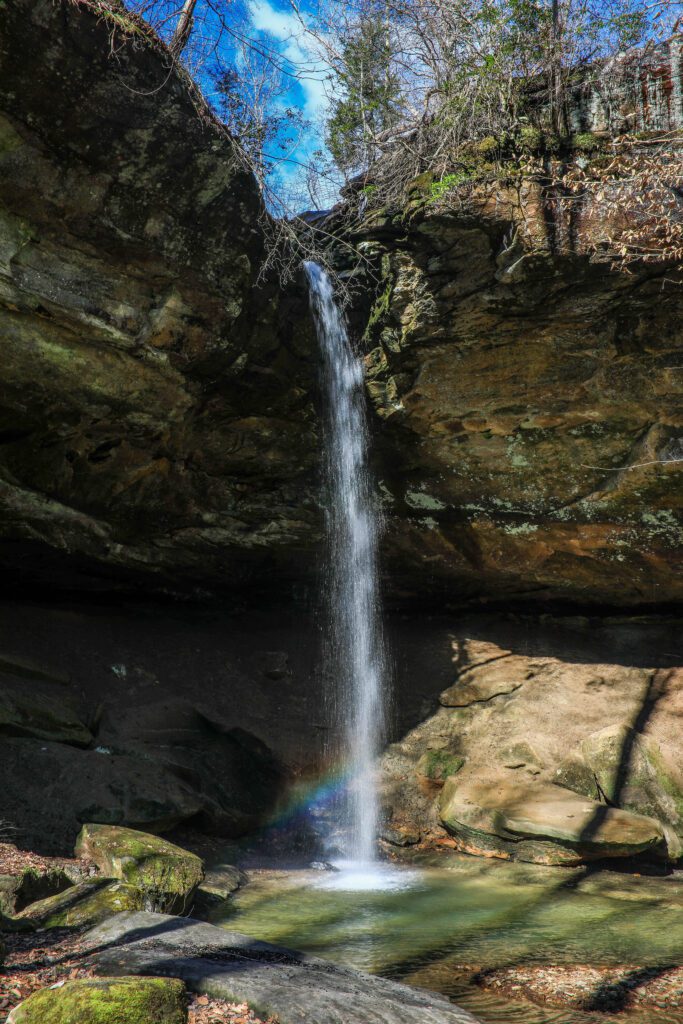 A narrow waterfall cascades down a rocky cliff into a small, clear pool below. Sunlight hits the water, creating a faint rainbow. Surrounding the waterfall are moss-covered rocks and trees with sparse foliage under a partly cloudy sky.