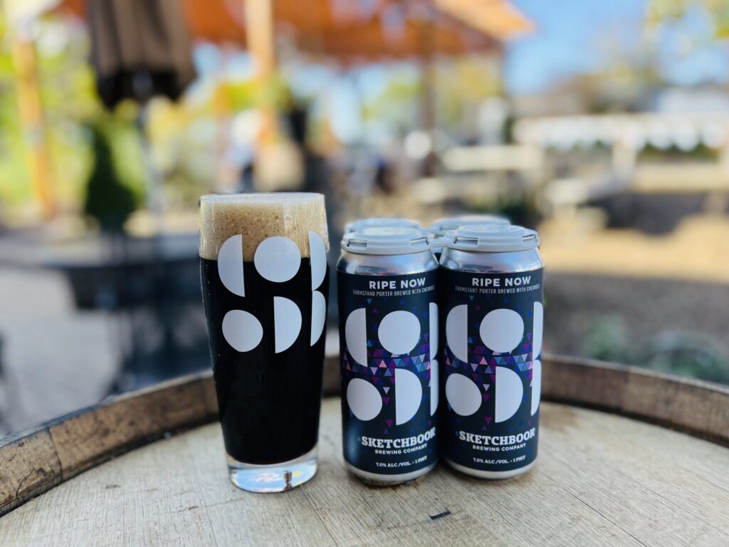 A pint glass filled with dark beer sits on a wooden barrel, next to a four-pack of cans marked Ripe Now by Sketchbook. The cans feature a modern design with abstract patterns. Blurred outdoor setting in the background.