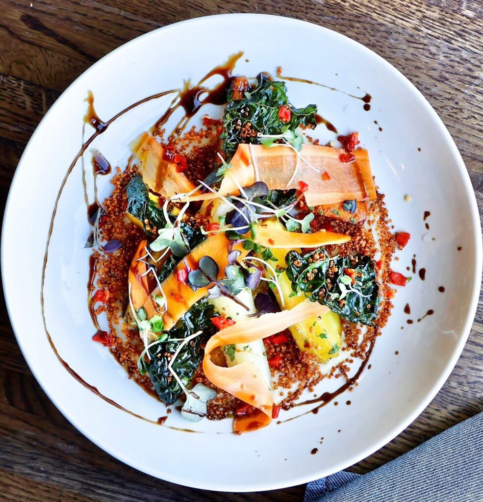 A white plate with a colorful dish arranged artistically. It features kale, thin carrot slices, sprouts, and quinoa, drizzled with a dark sauce. The dish is set on a wooden surface with part of a gray cloth visible.