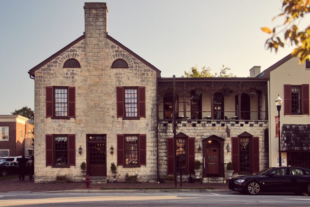 Historic two-story stone building with arched windows and wooden shutters. The left side has three windows, and the right has a decorative balcony. A black car is parked in front, with a classic streetlamp nearby.