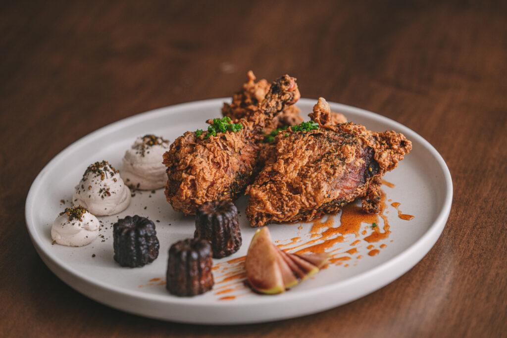 A white plate with crispy fried chicken drumsticks garnished with herbs, a sliced fig, small dark-colored cakes, and dollops of creamy sauce. Drizzles of sauce decorate the plate, set on a wooden table.