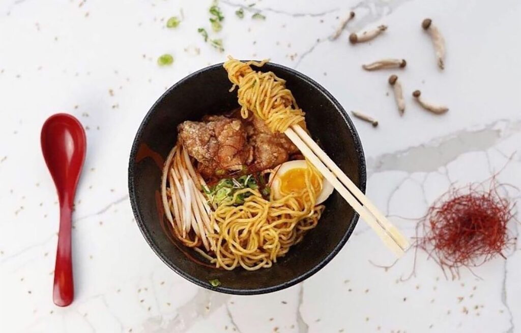 A black bowl of ramen on a marble surface, contains noodles, fried chicken, half a boiled egg, and scallions. Chopsticks rest on the bowl, with mushrooms and a red spoon nearby. Thin red seasoning strands are scattered around.