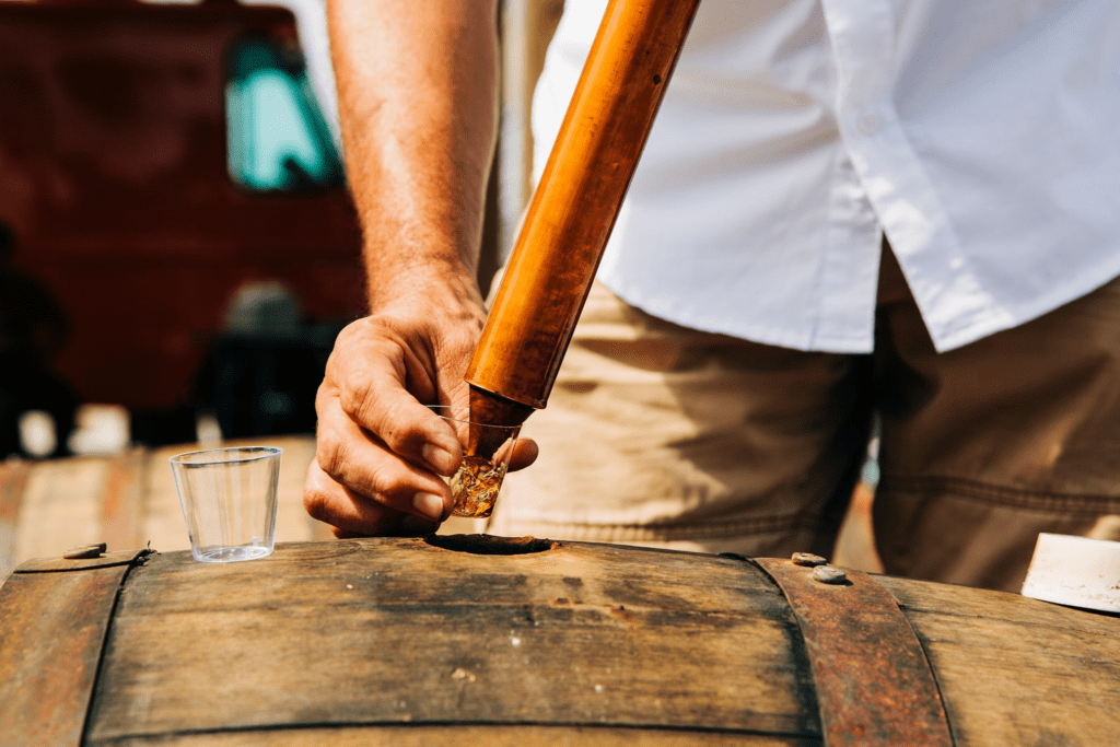 A person wearing a white shirt and khaki shorts uses a wooden tool to extract liquid from a barrel. A small glass sits nearby. The background is blurred, featuring a red item, possibly a vehicle.