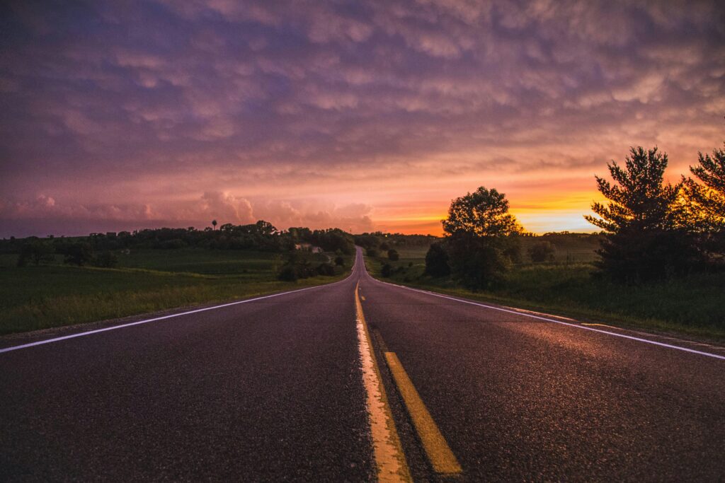 A rural road stretches into the distance under a dramatic sunset sky filled with purple and orange hues. Lush greenery lines the road, and trees are silhouetted against the vivid horizon.