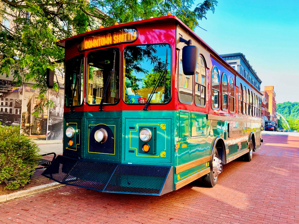 A vintage-style shuttle bus with Downtown Shuttle on the sign is parked on a brick road. The bus is green and maroon, with large windows. Trees and buildings are in the background under a clear blue sky.