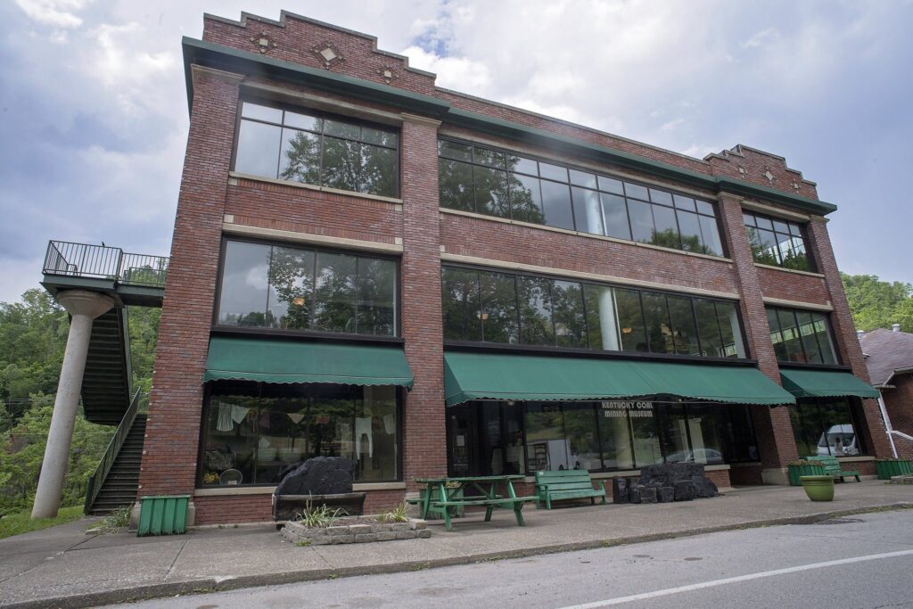 A three-story red brick building with large glass windows and green awnings, including a stairway on the left side. A green picnic table sits on the sidewalk in front, and trees are reflected in the windows.