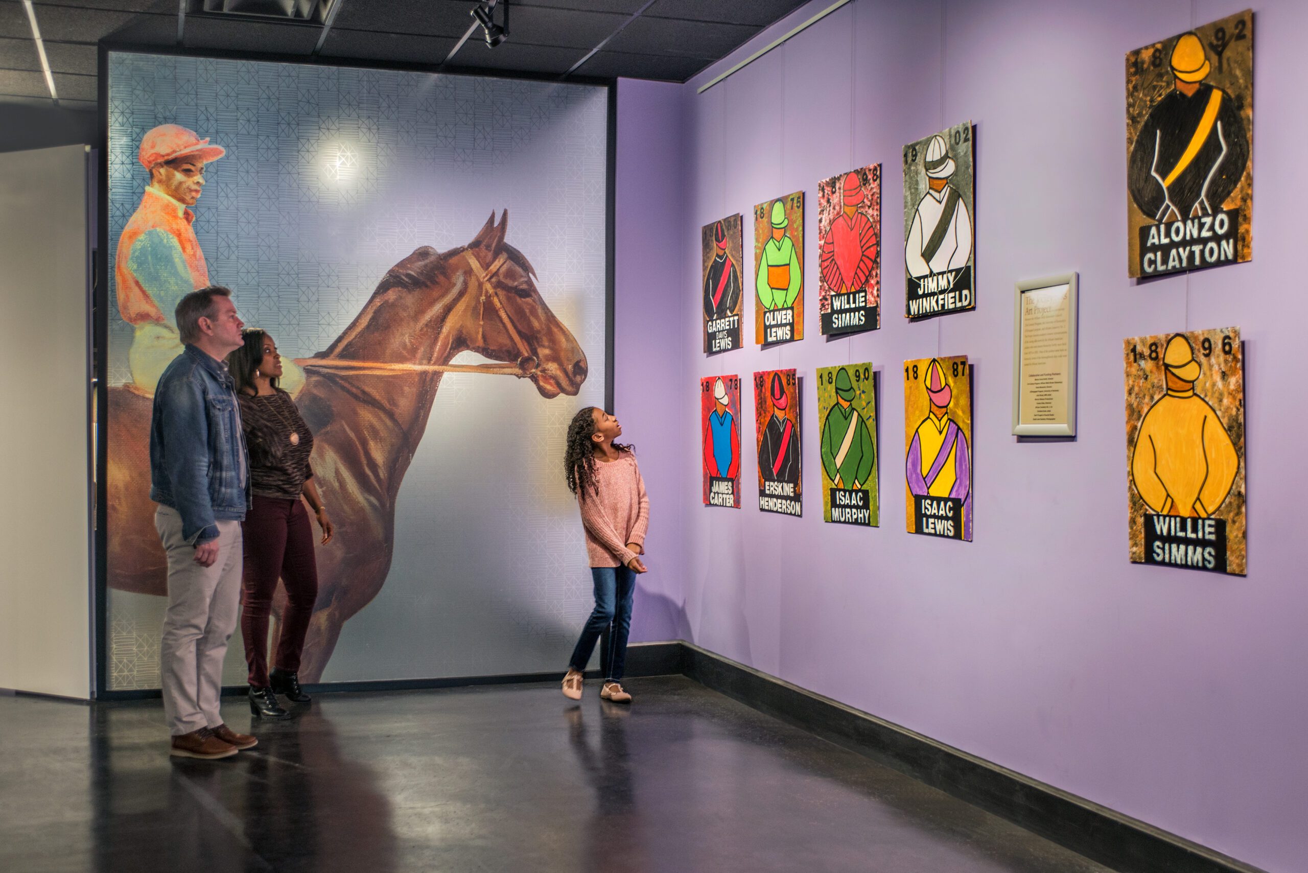 Three people view colorful jockey-themed artwork and portraits on a museum wall, with a large mural of a jockey on a horse in the background.