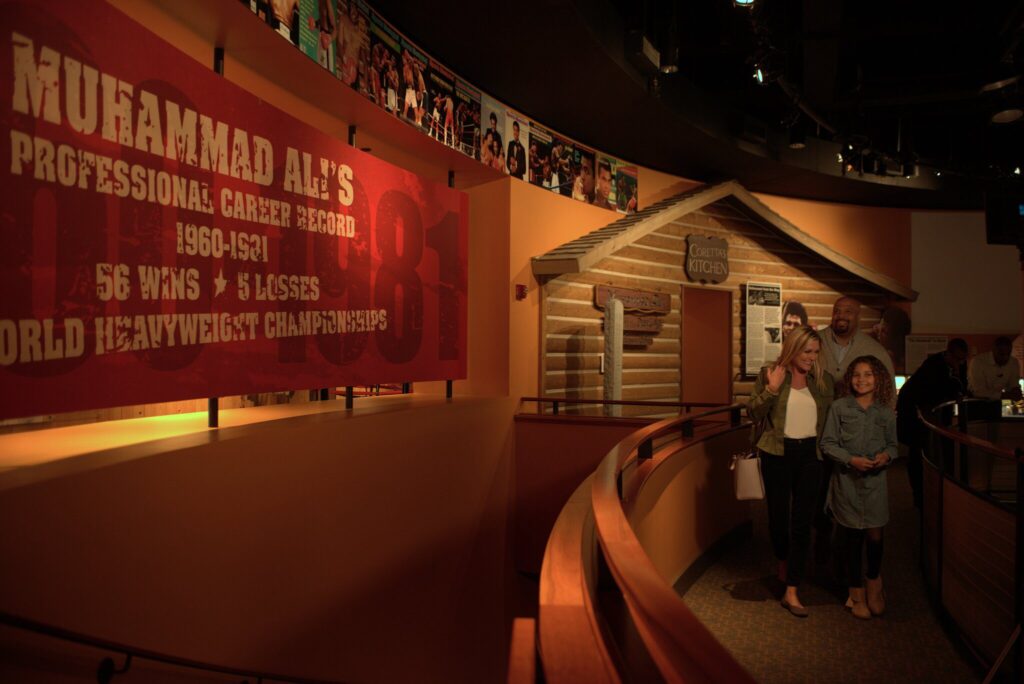 A family walks through a dimly lit museum exhibit featuring a large red sign about Muhammad Ali’s career and a replica of a rustic wooden house labeled “Carrie’s Kitchen.”.