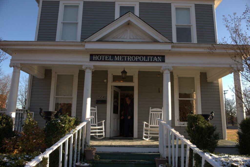 A gray, two-story house with white columns and trim, a sign reading Hotel Metropolitan above the door, and two white rocking chairs on the front porch. A person stands in the doorway.
