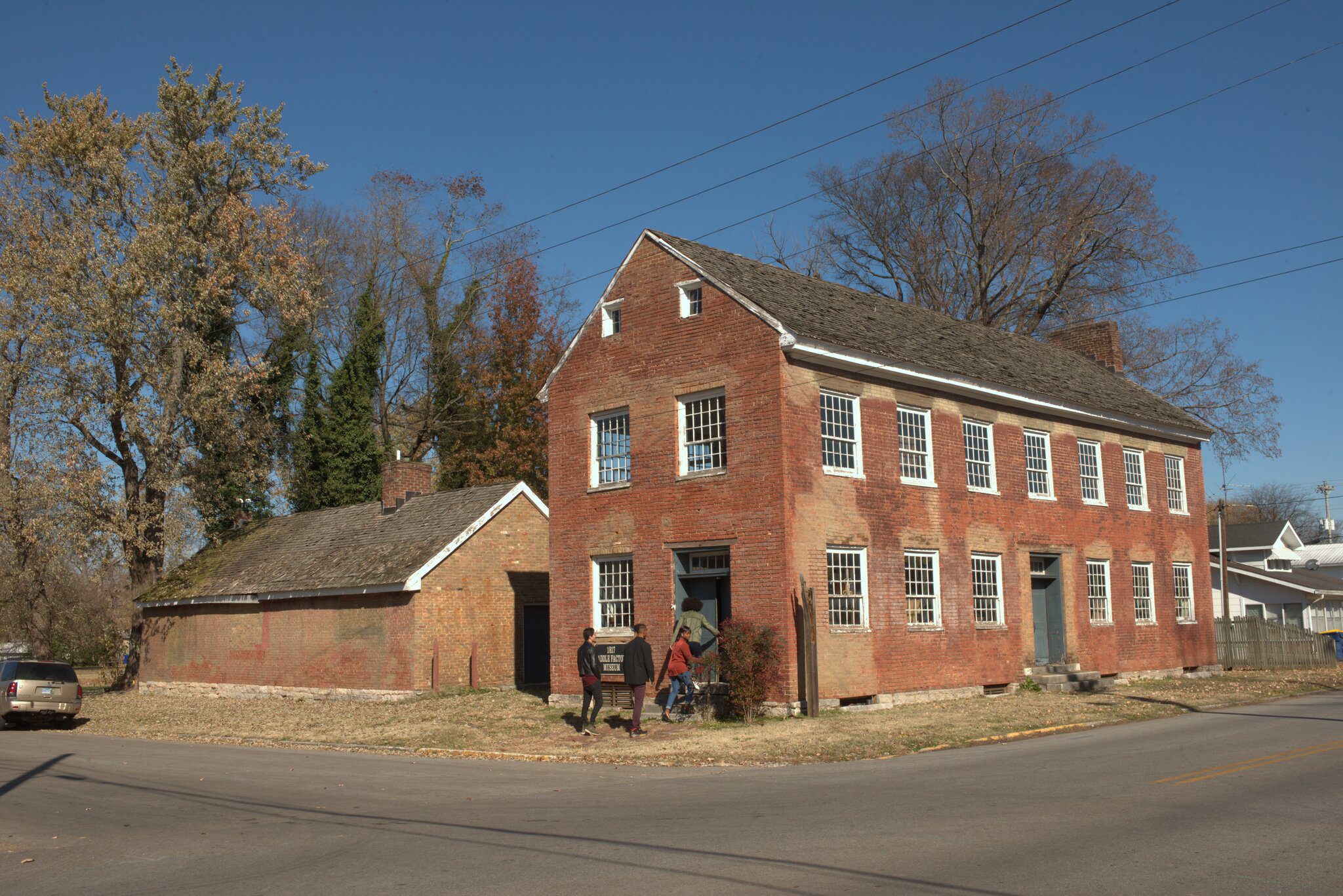 A historic two-story red brick building with white-trimmed windows stands on a street corner. A smaller brick structure is nearby, and two people walk toward a sign by the entrance under a clear blue sky.