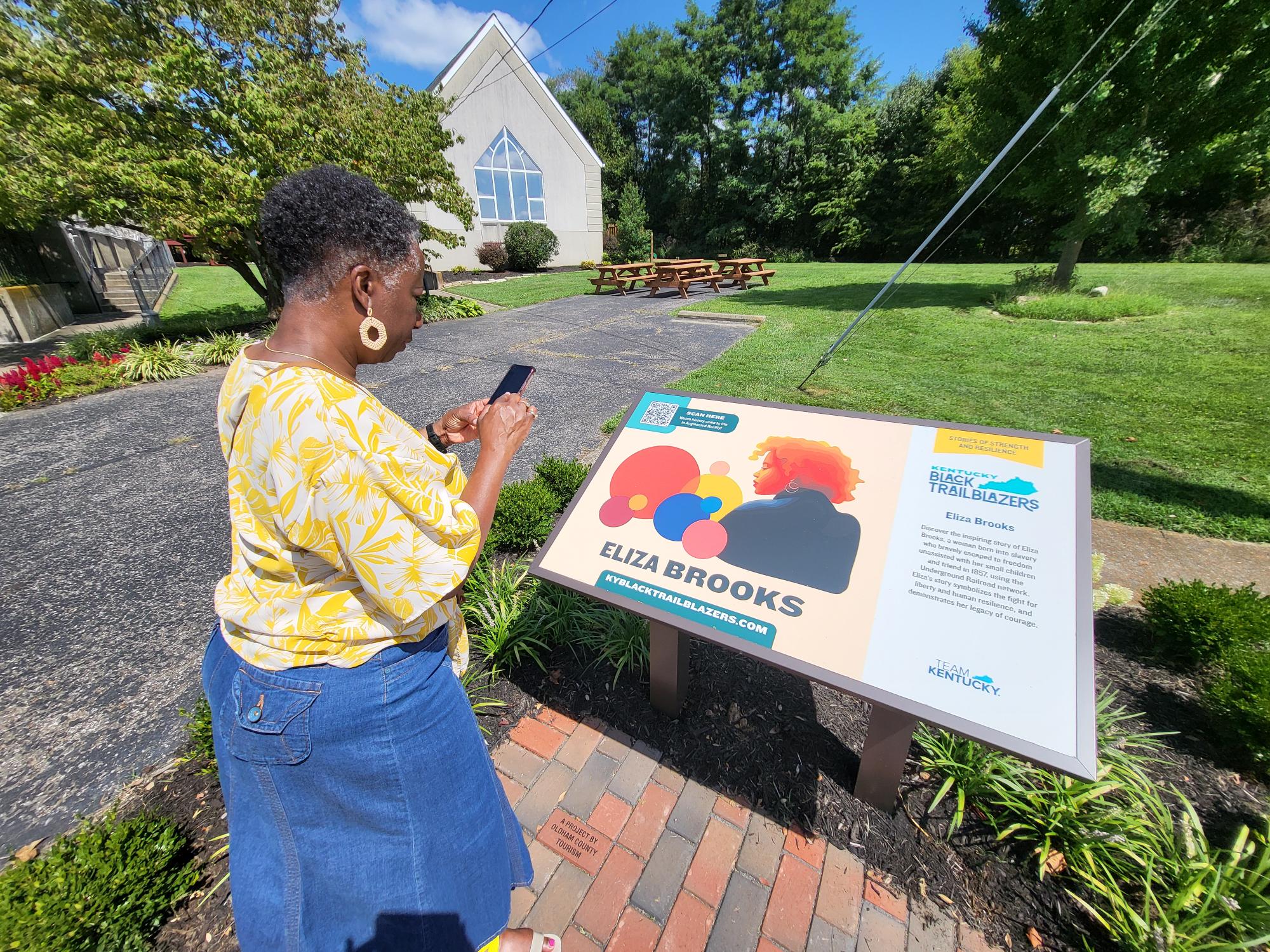 A woman in a yellow floral shirt views and takes a photo of an outdoor informational sign about Eliza Brooks, part of a Black Trailblazers exhibit, with a church and green trees in the background.