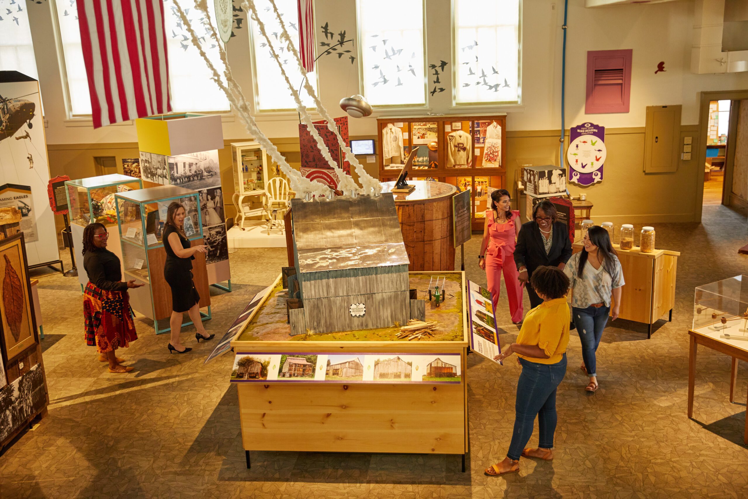 A group of people explore an indoor museum exhibit featuring model buildings, historical displays, and large photographs, with an American flag hanging in the background.