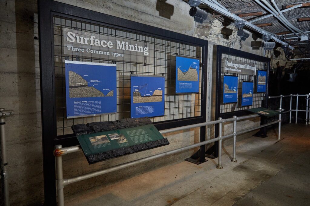 An indoor museum exhibit displays informational panels about surface and underground mining methods, with diagrams and text mounted on metal grids against a concrete wall. Metal railings separate the exhibit from the walkway.