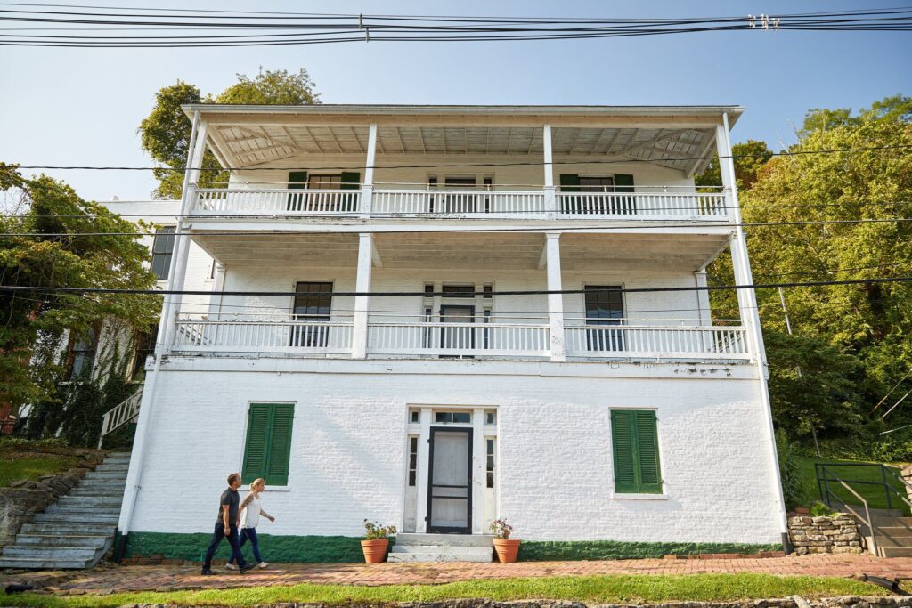 A large, three-story white house with green shutters and wide balconies on each floor. Two people walk on the sidewalk in front, with potted plants by the entrance and trees surrounding the house.