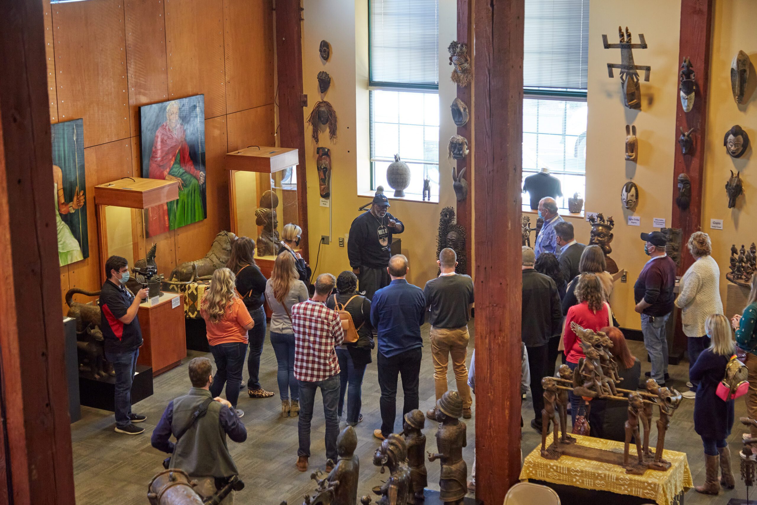 A group of people stand and listen to a tour guide inside a museum gallery with sculptures, masks, and paintings displayed on the walls and in glass cases. Natural light comes through large windows.