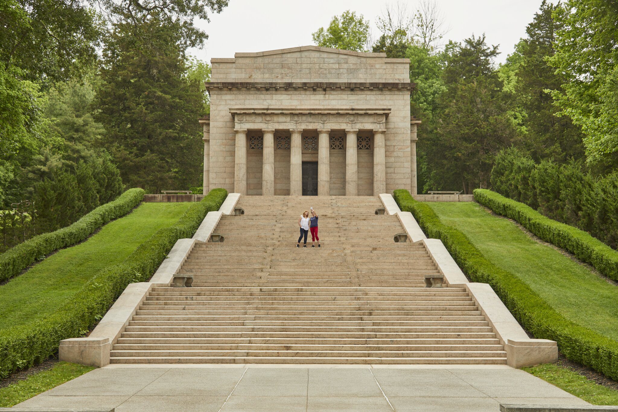 A wide stone staircase leads up to a large, classical-style memorial building surrounded by green trees and hedges. Three people stand near the top of the stairs in front of the entrance.