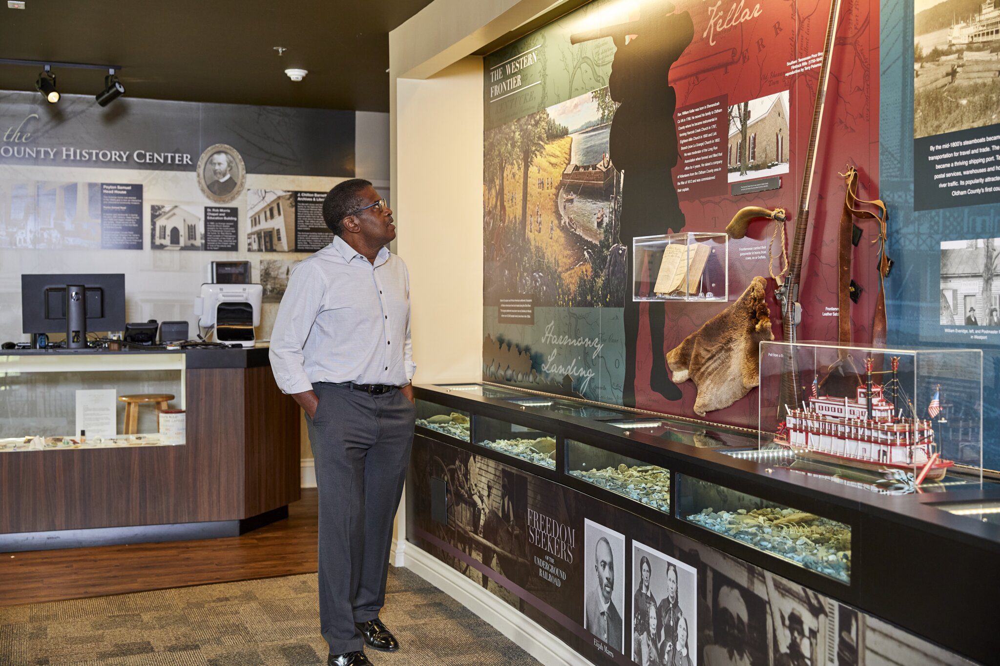 A man stands in a museum, looking at a display case with historical artifacts, photos, and informational panels. The background includes images, text, and a counter with a computer.