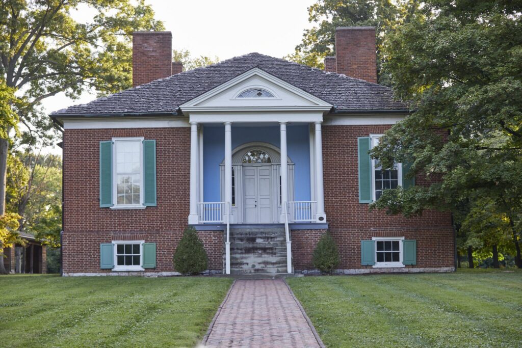 A historic brick house with green shutters, white columns, a central front door, and steps leading to the entrance, surrounded by trees and a lawn with a brick walkway.