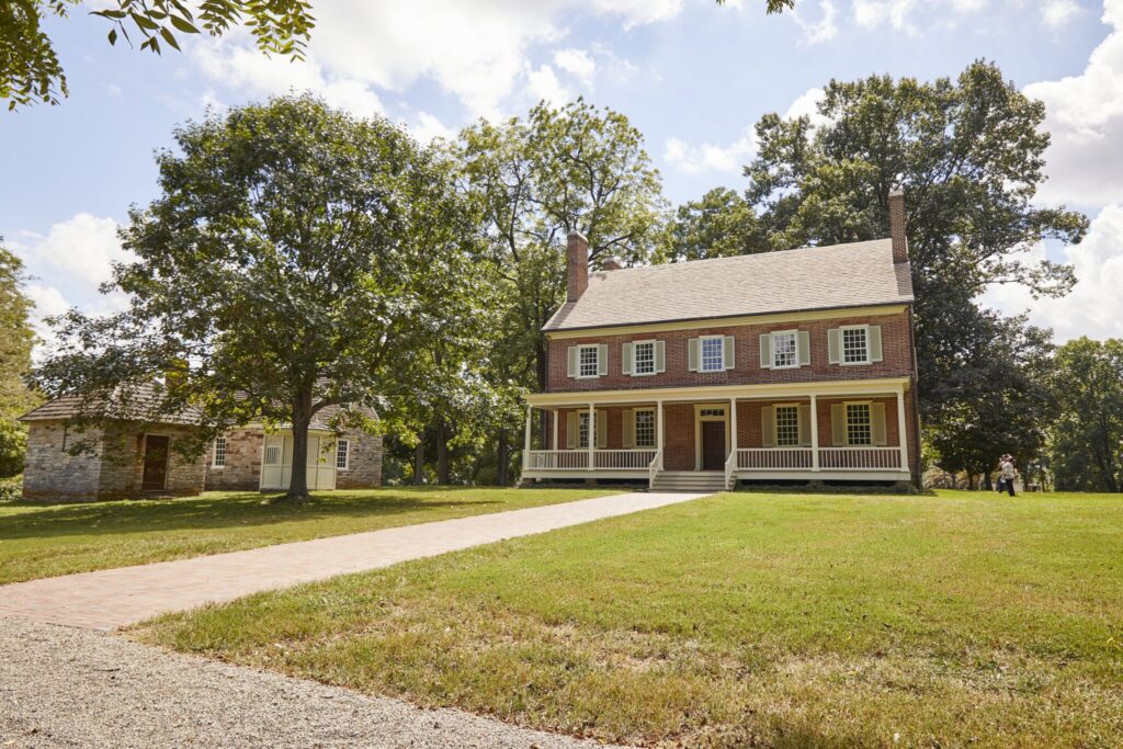 A two-story historic brick house with a front porch and white trim stands on a grassy lawn, surrounded by large trees and a smaller stone building to the left, under a partly cloudy sky.