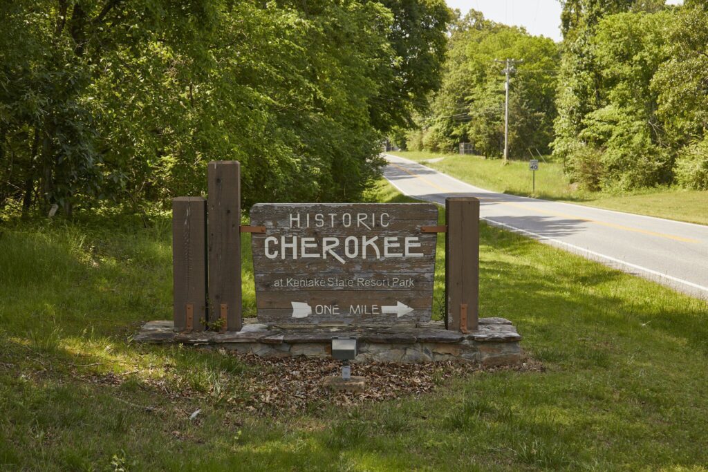 A wooden sign by a rural road reads Historic Cherokee at Kenlake State Resort Park, One Mile with an arrow pointing right. Lush green trees and grass surround the area under sunlight.