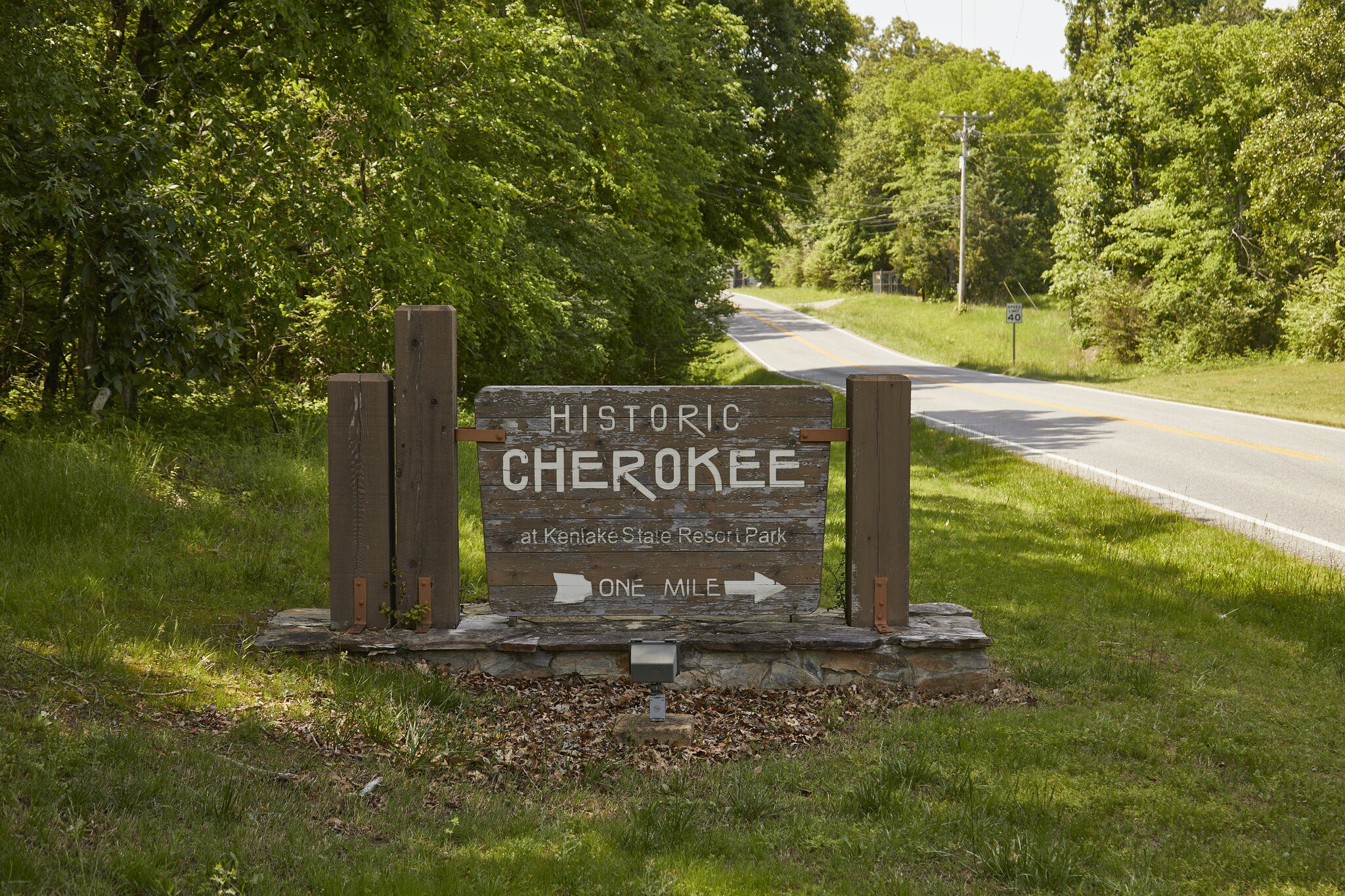 A wooden sign by a rural road reads Historic Cherokee at Kenlake State Resort Park, One Mile with an arrow pointing right. Lush green trees and grass surround the area under sunlight.