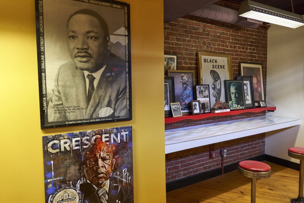 A room with exposed brick walls features framed photos and posters of Black historical figures, including Martin Luther King Jr., displayed above a counter with red stools beneath a shelf lined with more portraits.