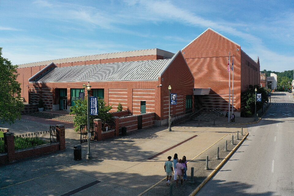 A large brick building with green doors, steps leading to multiple entrances, and two flagpoles out front. Four people walk on the sidewalk nearby under a clear blue sky.