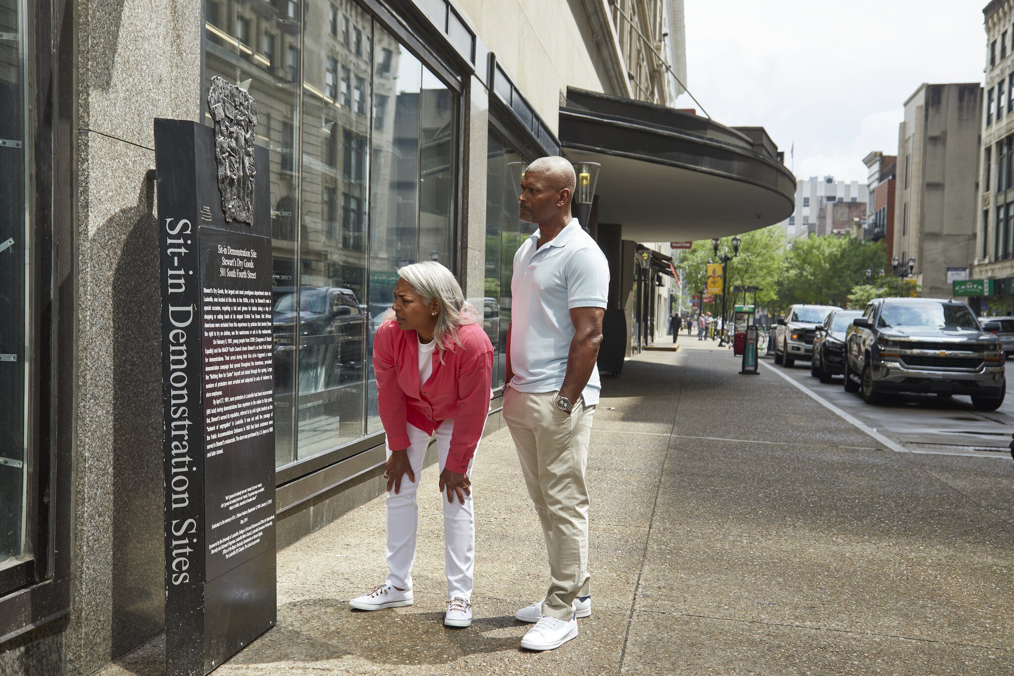 An older woman and a man stand on a city sidewalk, reading a historical marker titled Sit-in Demonstration Sites in front of a glass storefront. Cars are parked along the street in the background.
