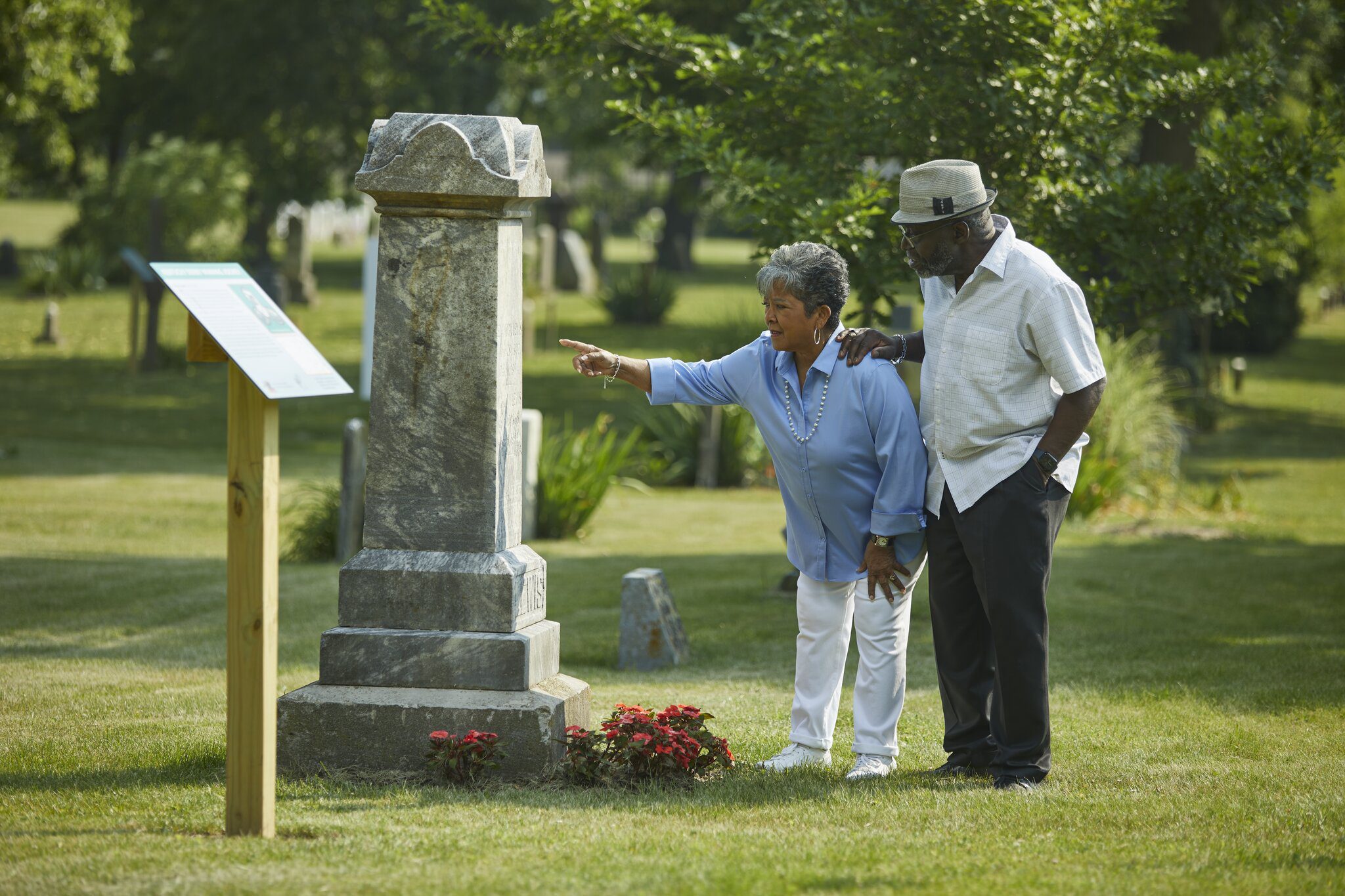 An older couple stands beside a gravestone in a cemetery. The woman is pointing at an informational sign, while the man stands next to her with his hand on her shoulder. Lush green trees and grass surround them.