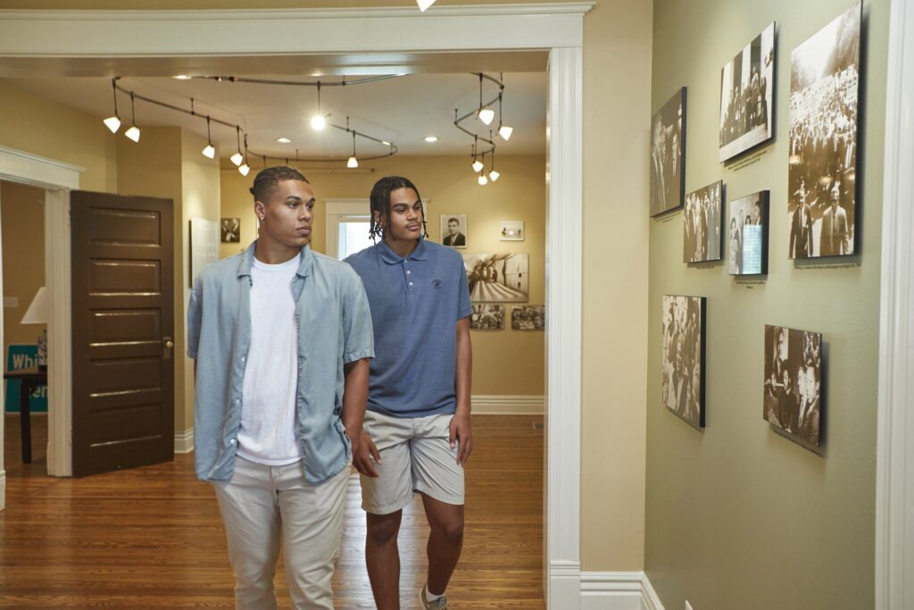 Two young men walk through a hallway with wooden floors and cream-colored walls, looking at black-and-white photographs displayed on the wall. The space is well-lit with modern ceiling lights.