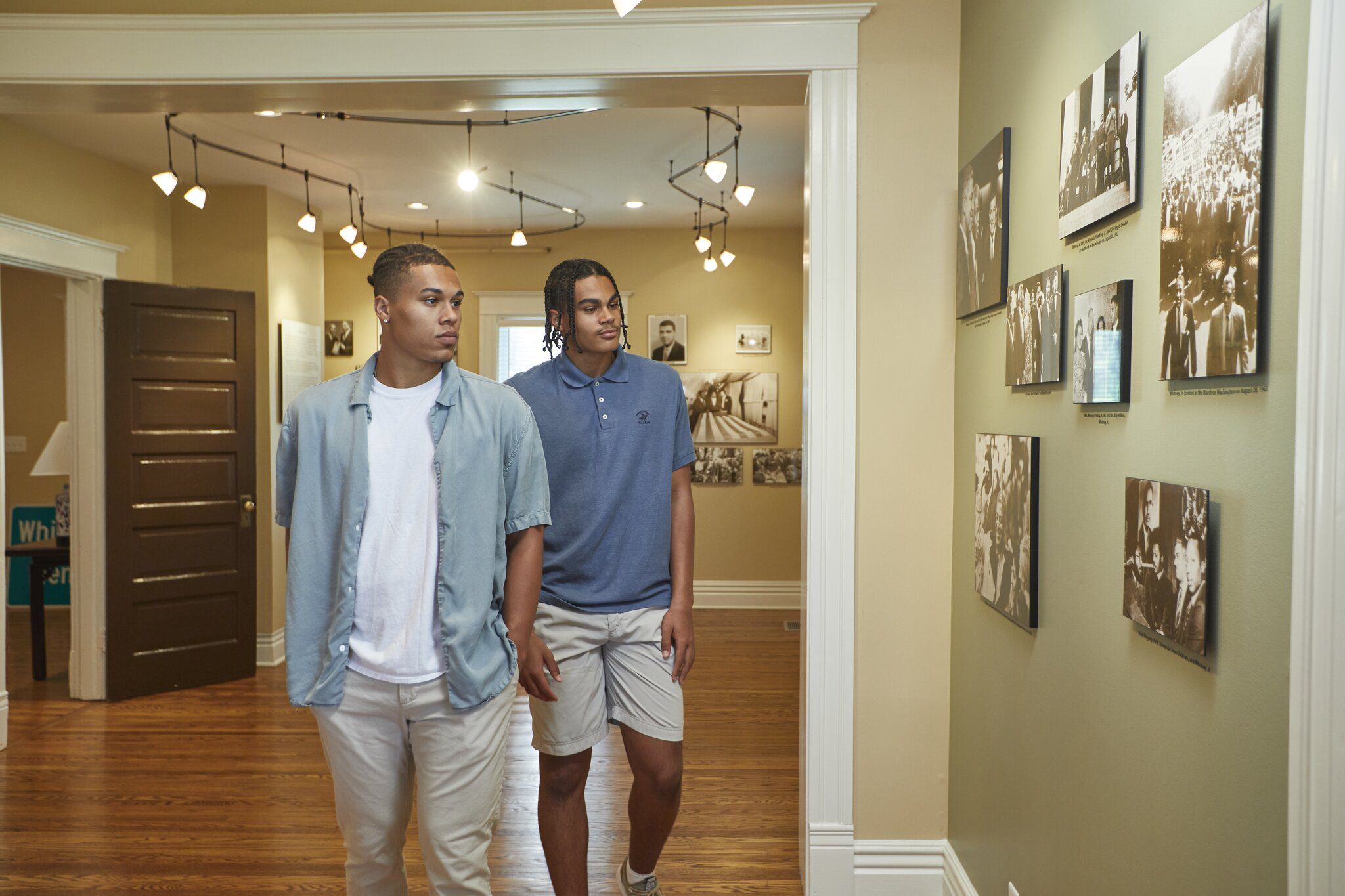 Two young men walk through a hallway with wooden floors and cream-colored walls, looking at black-and-white photographs displayed on the wall. The space is well-lit with modern ceiling lights.