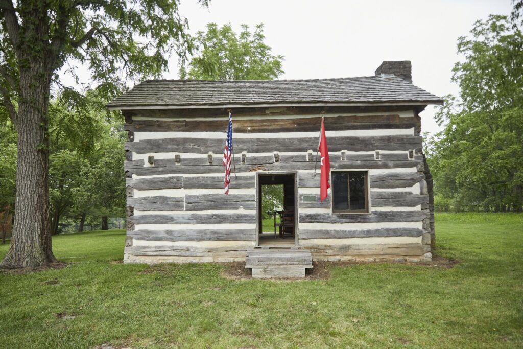A small, historic log cabin with two flags—one American and one red—hanging beside the entrance, surrounded by green grass and trees. The cabin has a single open door and one visible window.