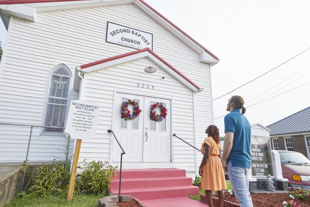 A man and a woman stand outside a white church with red trim, looking at its entrance decorated with two wreaths. The sign above the doors reads “Second Baptist Church.”.