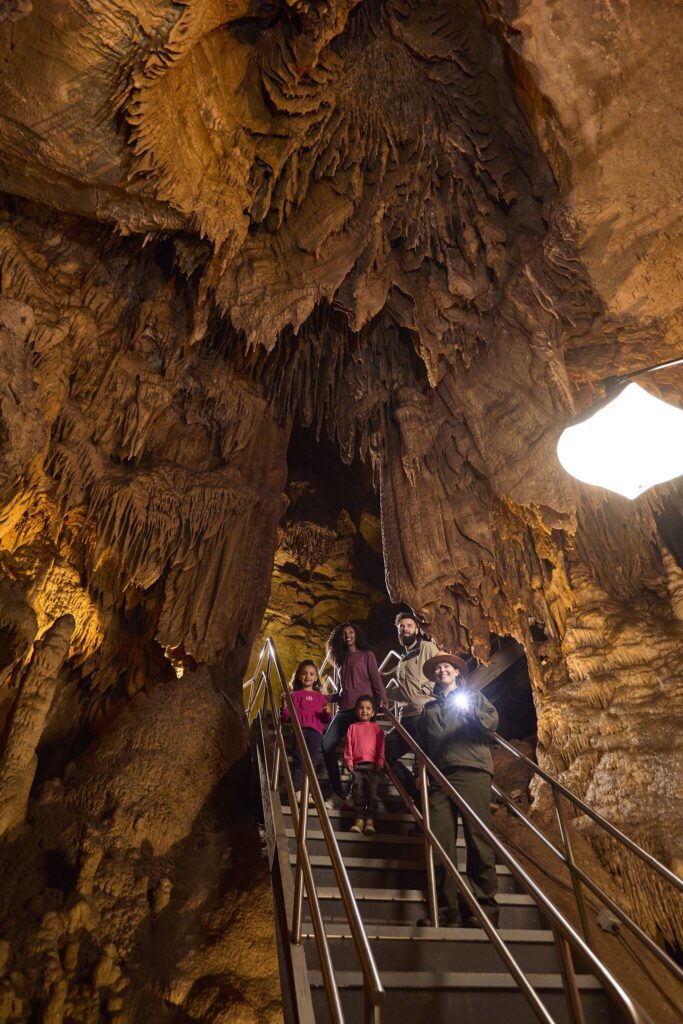 A group of people stands on a metal staircase inside a cave, surrounded by large, jagged stalactites and rock formations, illuminated by artificial lights.