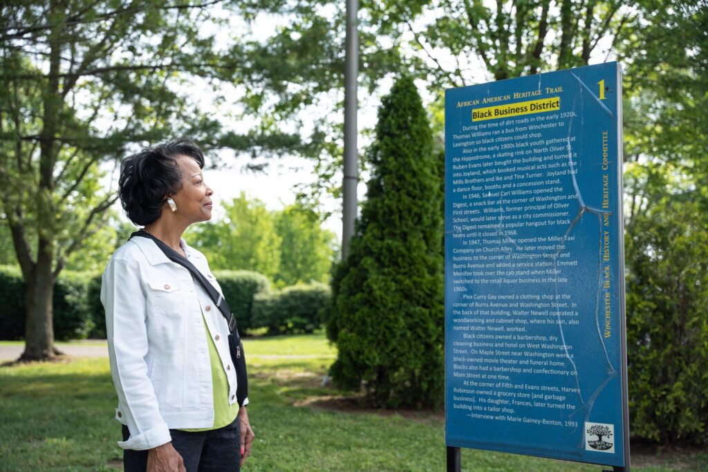 A woman wearing a white jacket stands outdoors, reading an informational sign about the Black Business District on the American American Heritage Trail. Green trees and grass surround her in the background.