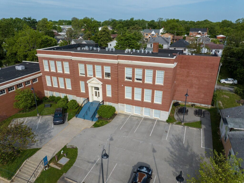 Aerial view of a red brick, three-story school building with large white-framed windows, a pedimented entrance, and a small parking lot with cars in front. Green trees and residential houses surround the property.